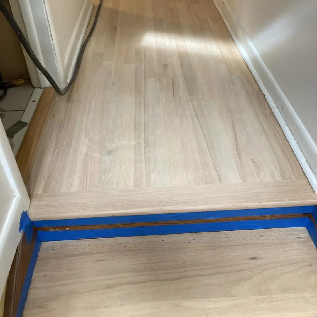 Freshly sanded light hardwood floor in a hallway with blue tape along a step